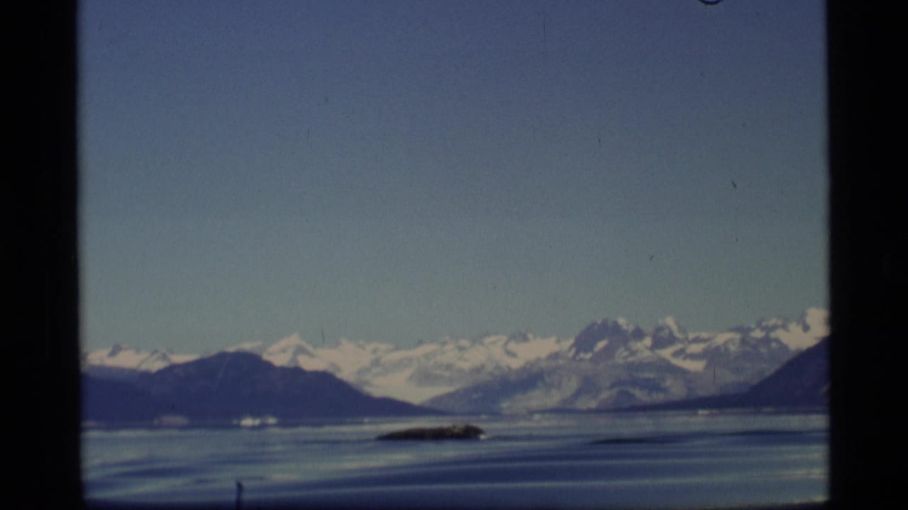 Snow-capped mountains beyond calm blue water seen from an aircraft.