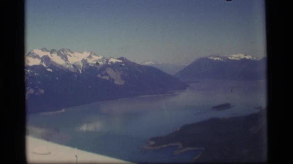 1977 Alaska aerial footage looking over a broad inlet, dark mountain slopes, snow patches, and the edge of an aircraft wing.