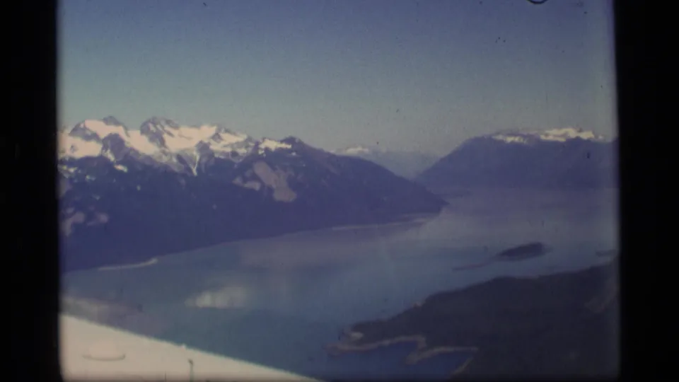 1977 Alaska 8mm frame showing a long blue waterway between steep mountain walls, with snow on higher peaks and aircraft framing below.