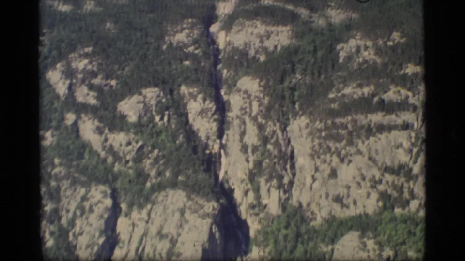 1977 Alaska aerial view of rugged rock formations and forested slopes, captured with soft 8mm grain and slight motion blur.