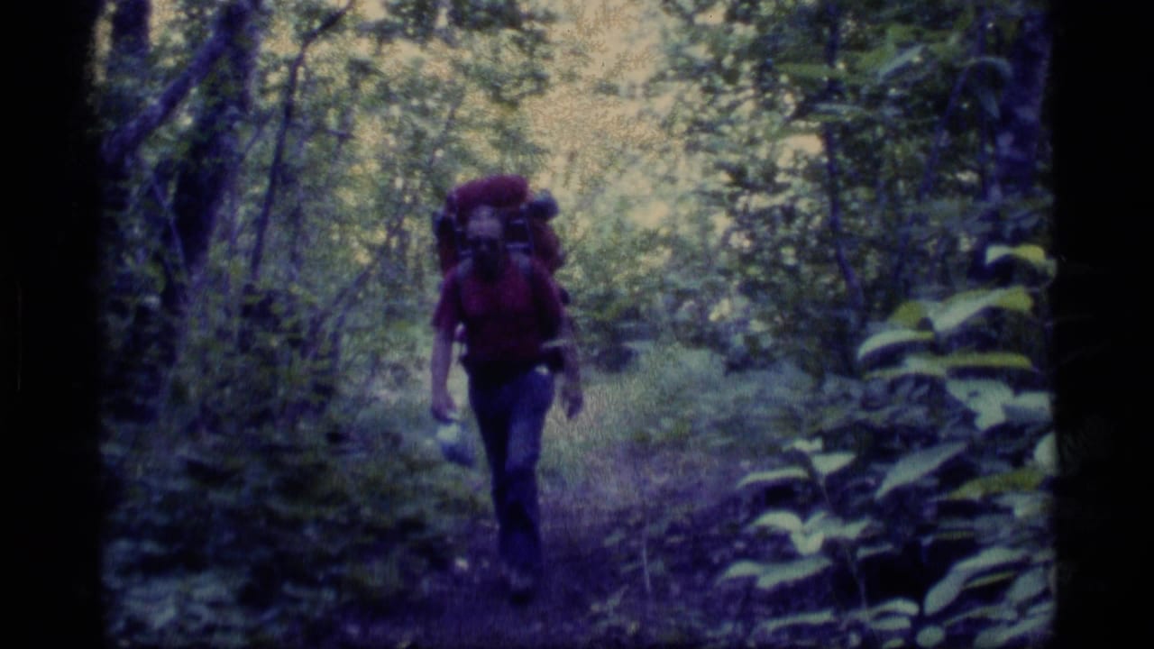 In Alaska 1977 home-movie footage, an adult hiker in a red shirt approaches through dense forest carrying a large backpack.