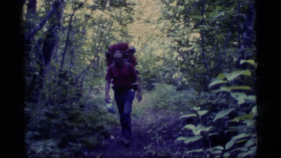 In Alaska 1977 home-movie footage, an adult hiker in a red shirt approaches through dense forest carrying a large backpack.