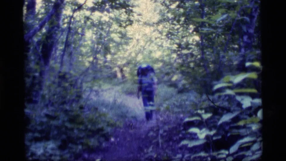 A second backpacker appears farther along the forest path in the 1977 Alaska home-movie sequence, surrounded by trees and brush.