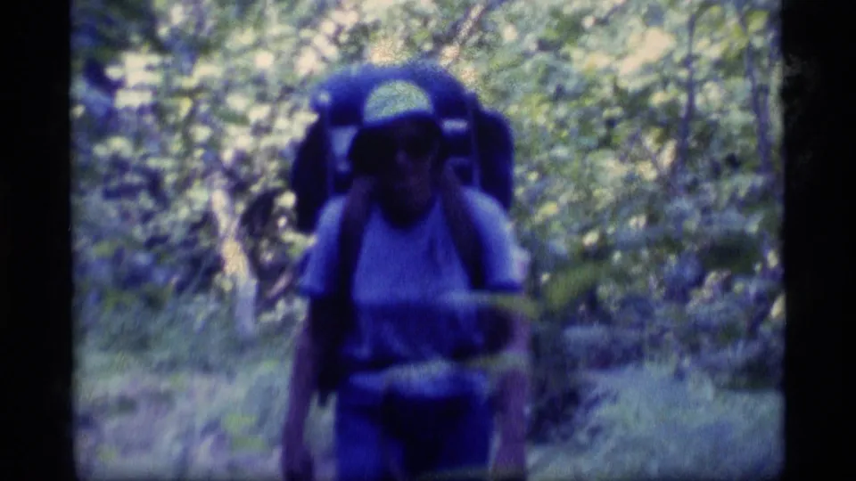 A backpacker wearing a light shirt and white cap approaches the camera on the Alaskan forest trail, carrying a large external-frame pack.