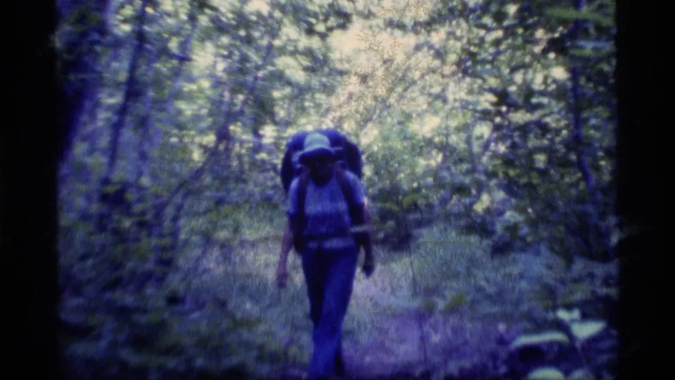 A backpacked hiker walks away through dense Alaska woodland in 1977 home-movie footage with a blue-purple film cast.