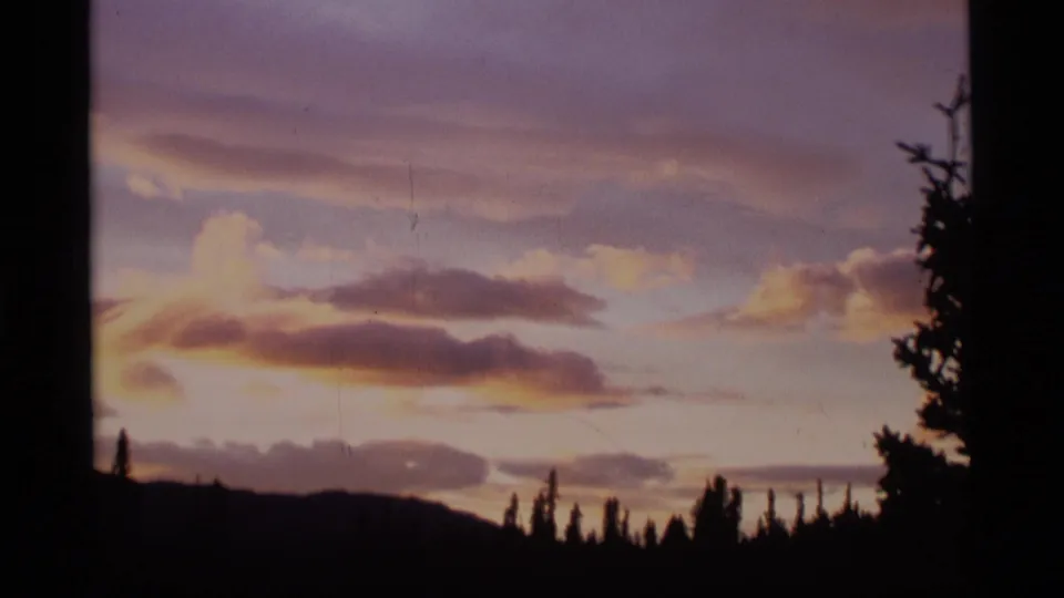 1977 Alaska 8mm frame showing purple and gold sunset clouds above silhouetted conifers and low hills.