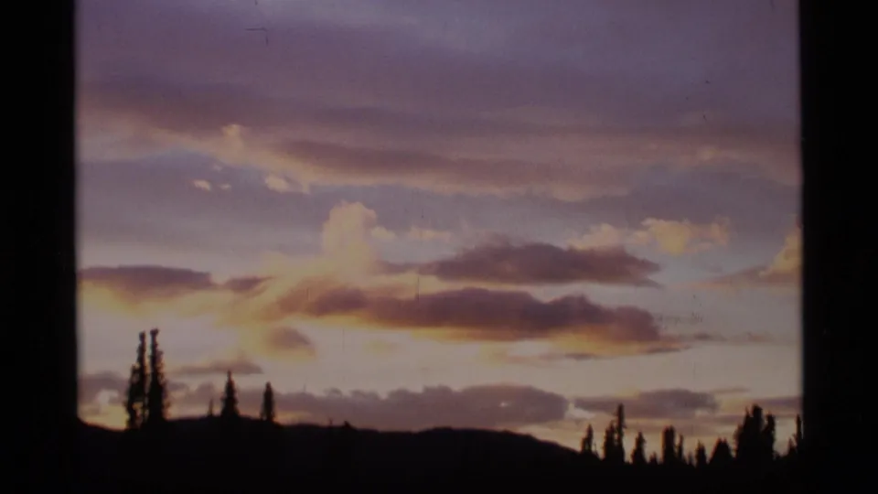 1977 Alaska home-movie frame with layered sunset clouds and a dark conifer skyline beneath the fading light.