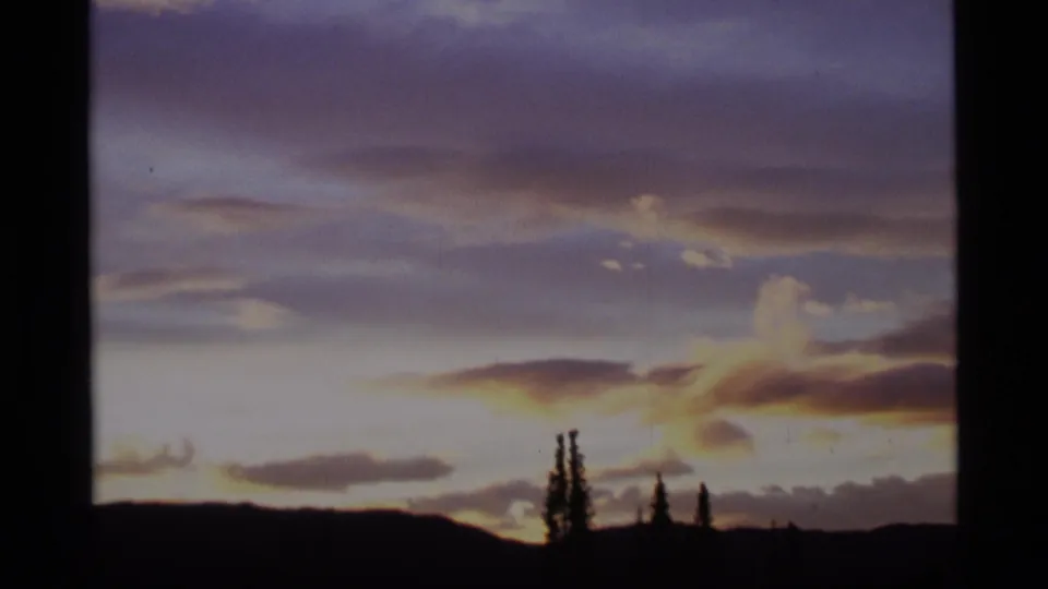 1977 Alaska home-movie frame showing a dimming sunset sky above dark hills and isolated conifer silhouettes.