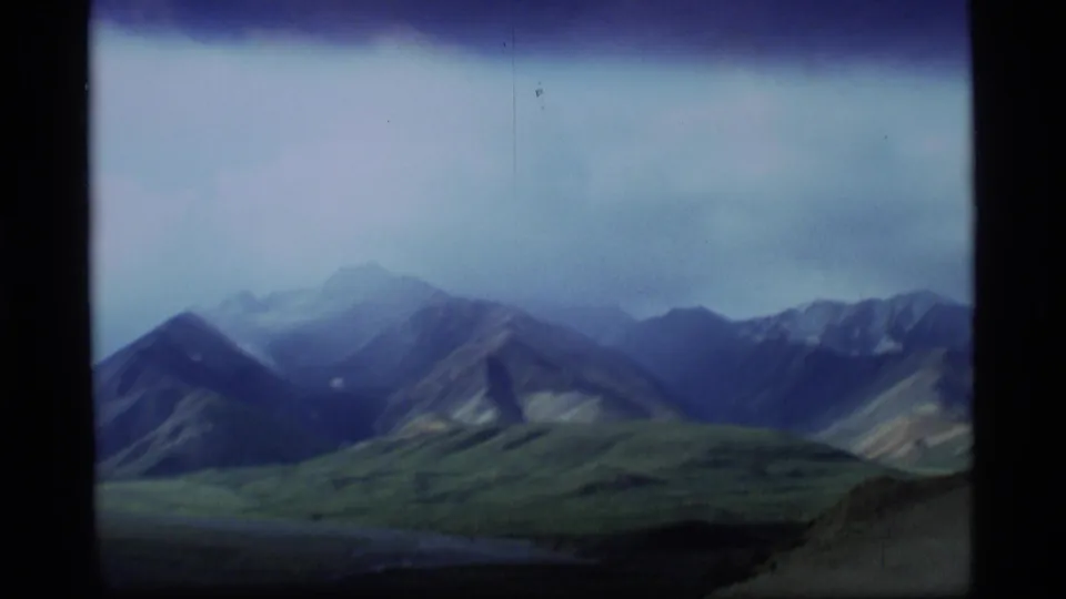 1977 Alaska 8mm frame showing distant rugged mountains and green valley terrain beneath a pale overcast sky.