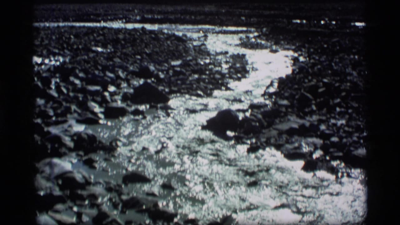 1977 Alaska 8mm frame of a shallow rocky stream with bright reflected light across water and stones.