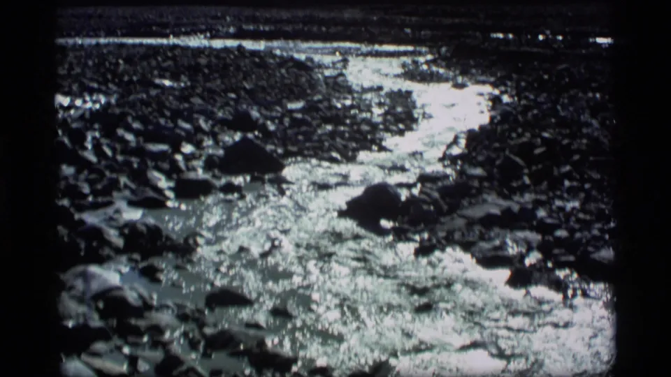 1977 Alaska 8mm frame of a shallow rocky stream with bright reflected light across water and stones.