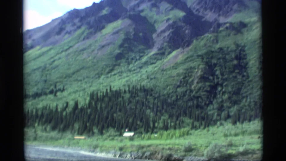 1977 Alaska home-movie frame of green mountain slopes, dense conifer forest, and a small distant structure near the base.