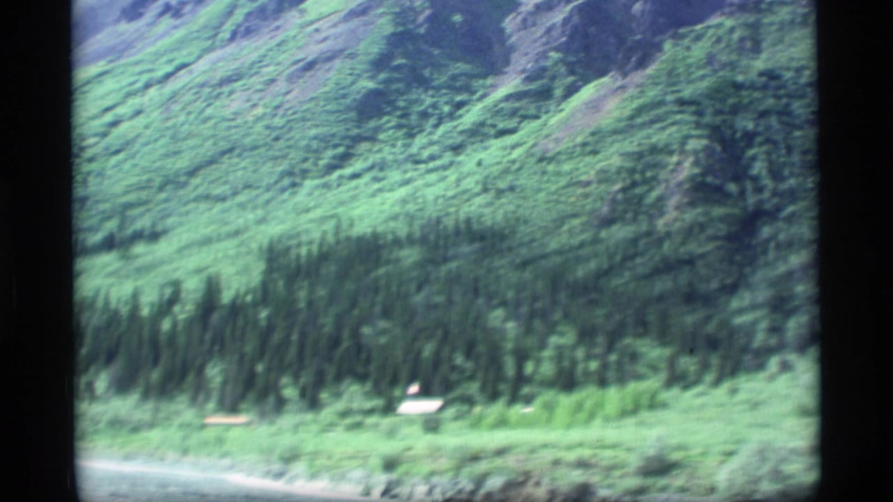 Green mountain slope with evergreen trees and small pale structures near the lower edge.