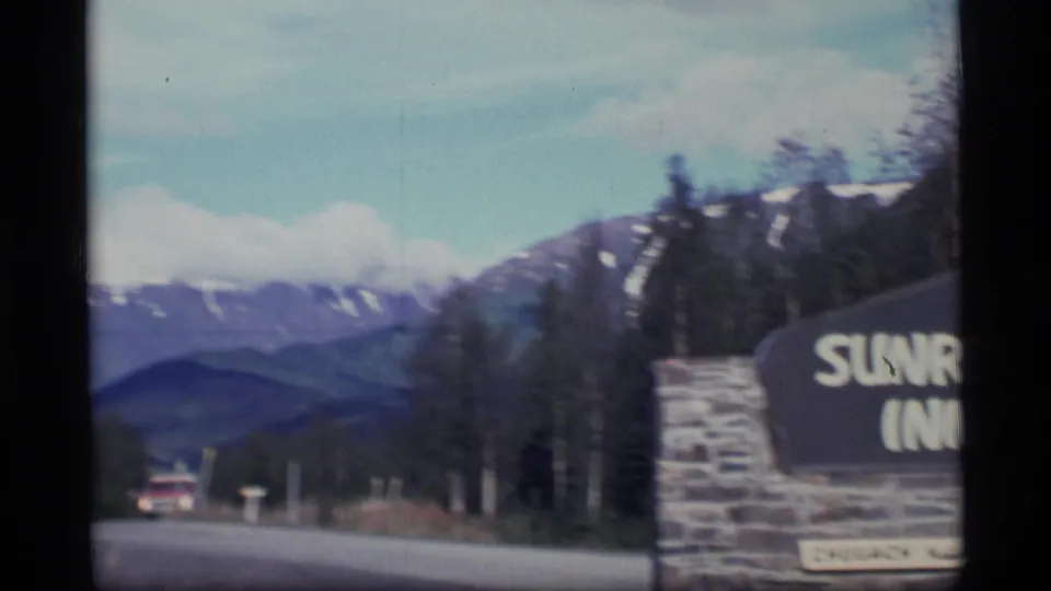 Roadside view in Alaska showing a stone Sunrise Inn sign beside a road with snow-capped mountains in the distance.