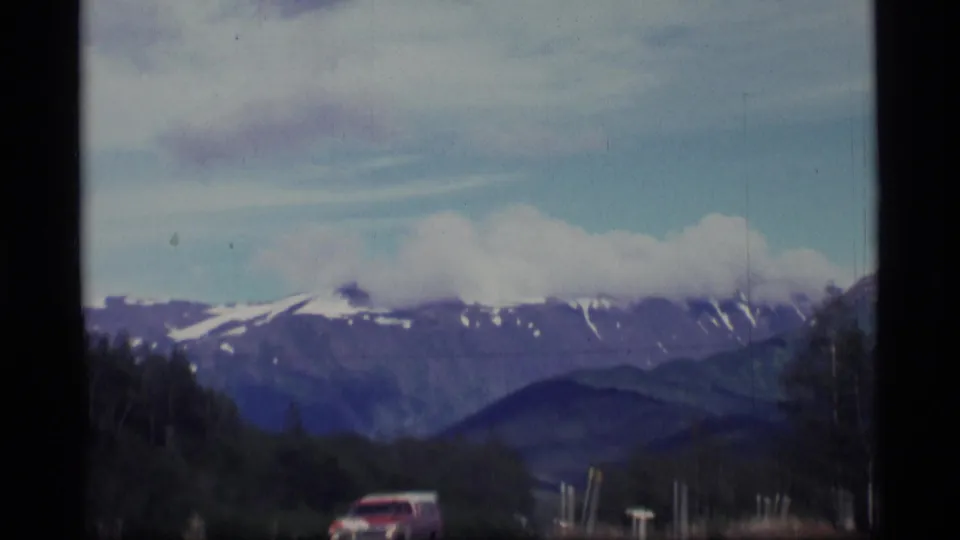 A red vehicle travels along a road through an Alaskan mountain landscape, with forest and snow-capped peaks beyond.
