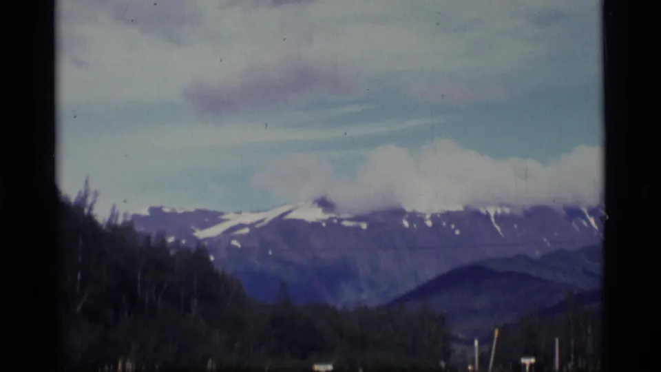 Snow-capped Alaskan mountains rise beyond forest and roadside posts in a soft, grainy 8mm travel frame.