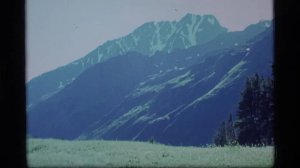 1977 Alaska 8mm landscape frame showing a grassy meadow, dark mountain slopes, snow-streaked peaks, and trees at the edge.
