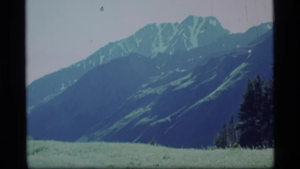 1977 Alaska mountain landscape with a broad meadow foreground, dark trees, and visible 8mm film border.
