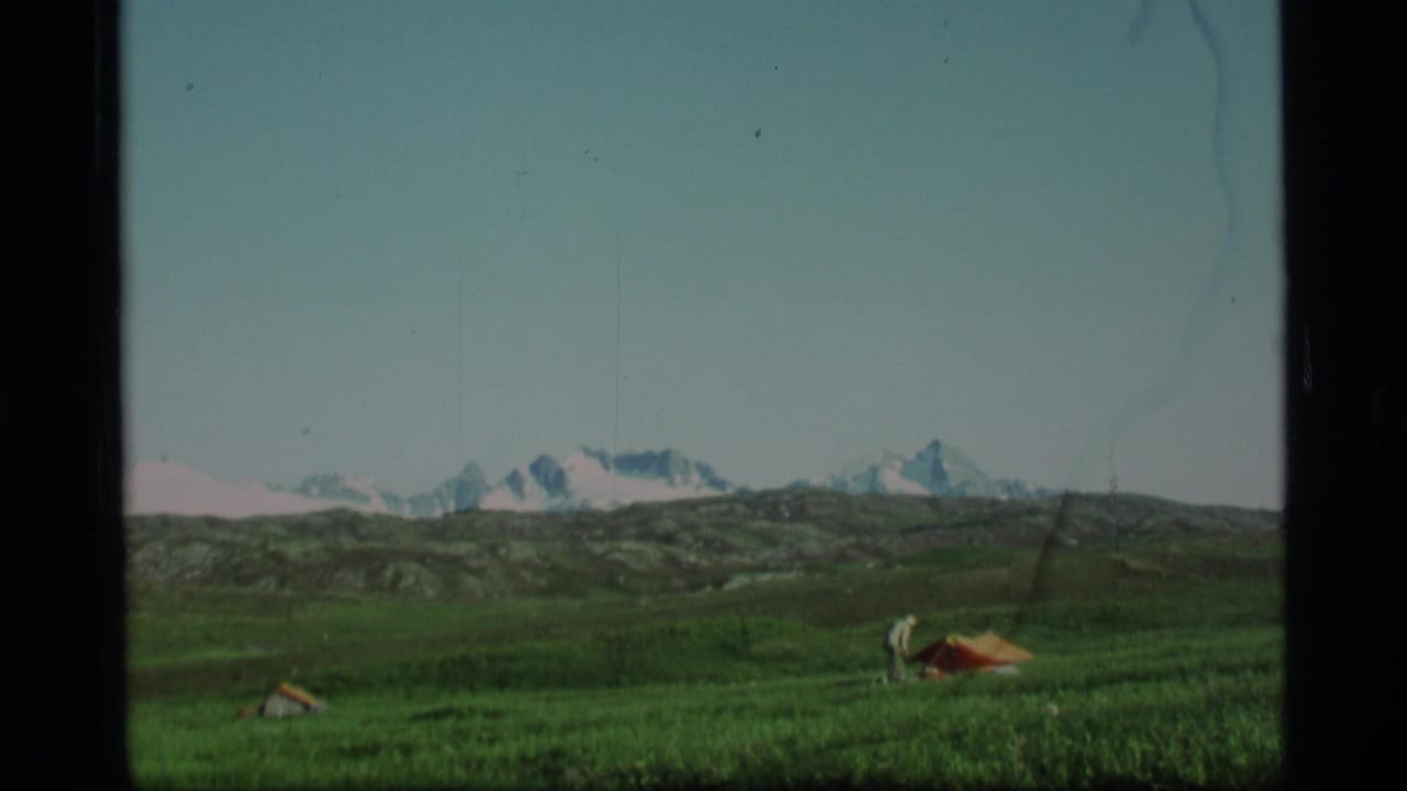 Green alpine meadow with small tents and snowy mountains in the distance.