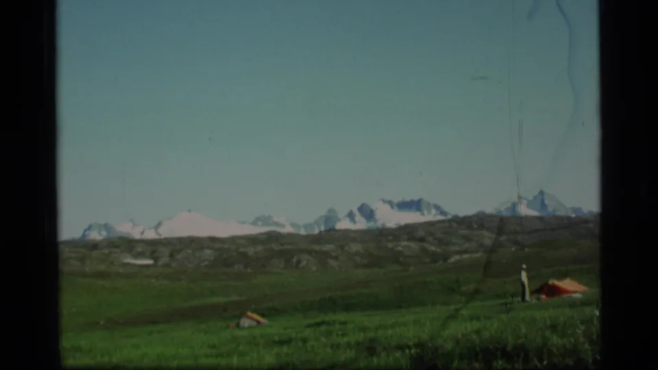 1977 Alaska 8mm frame with green open ground, low hills, snowy peaks, and a small orange tent near the right edge.