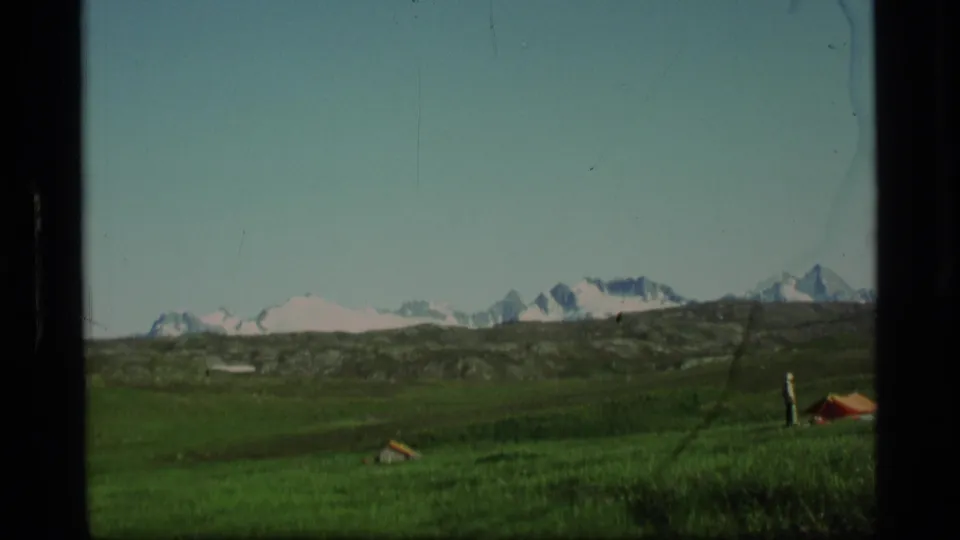 1977 Alaska 8mm frame showing a standing camper near a small tent in an alpine meadow below snowy mountains.