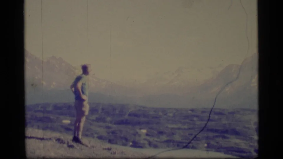 1977 Alaska 8mm frame showing a lone person in profile on rocky terrain with hazy snow-covered mountains in the background.
