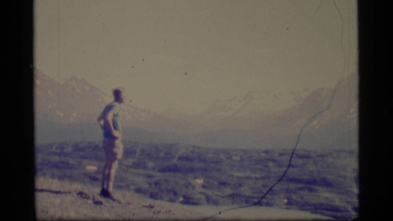1977 Alaska 8mm frame showing a man in light clothing standing on a rocky overlook above a broad mountain valley.