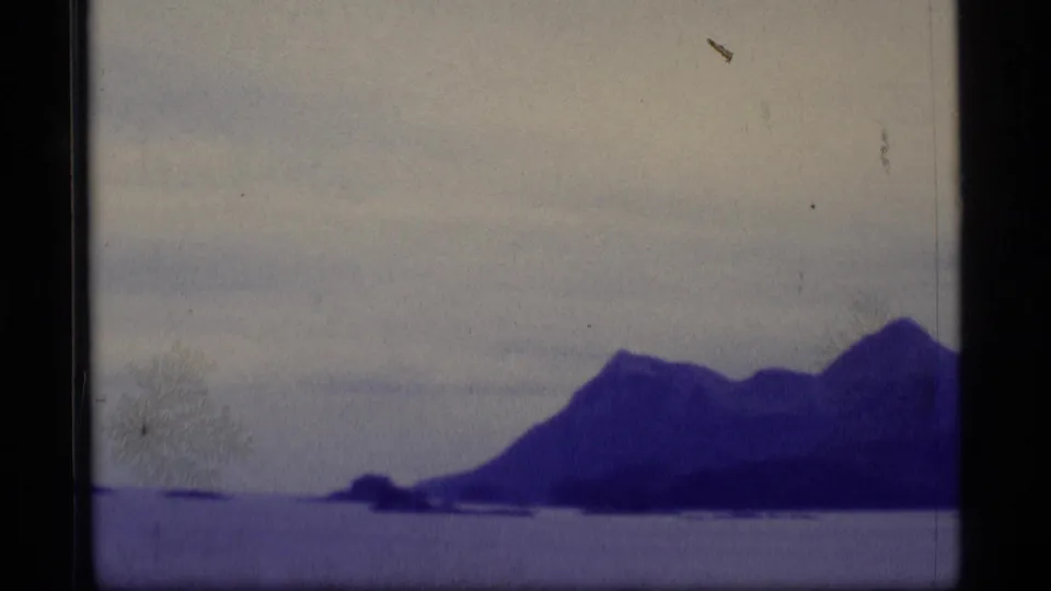 Purple-blue 8mm frame of an Alaskan mountain shoreline across calm water, with overcast sky and visible film wear.