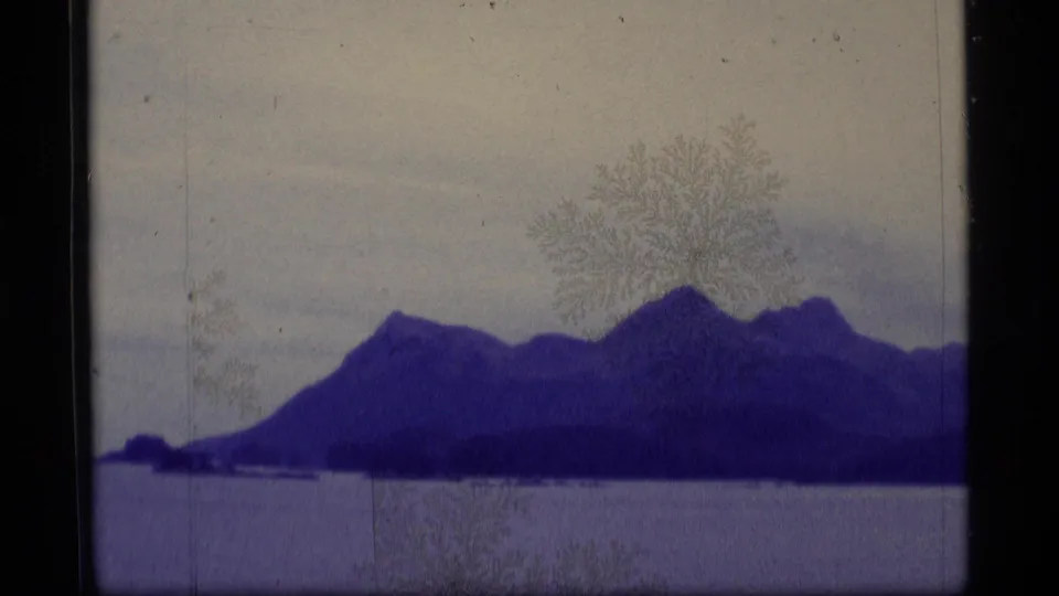 1977 Alaska 8mm frame showing a purple-tinted mountain range beyond water, with soft focus, film dust, and a pale sky.