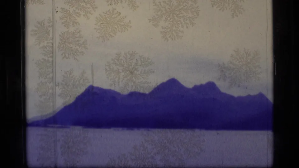 1977 Alaska home-movie frame showing a dark mountain shoreline across still water with soft grain and visible film border.