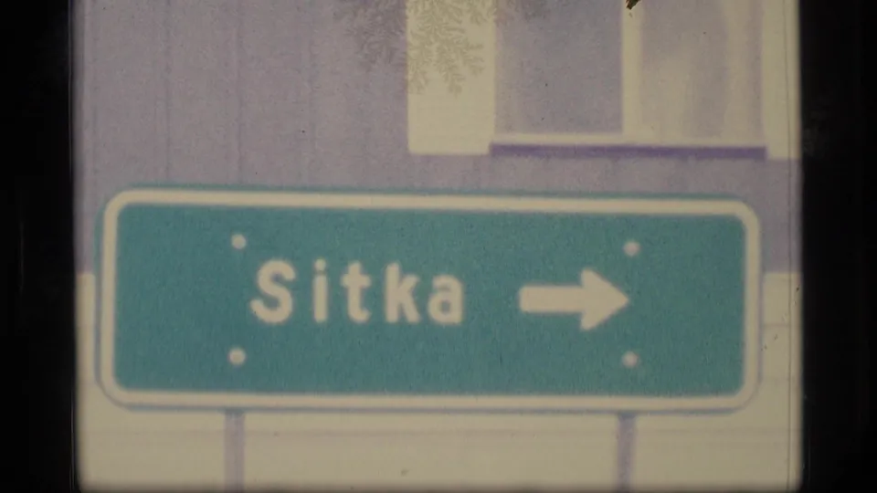 1977 Alaska frame with a green Sitka direction sign and right arrow, filmed close beside light building siding and windows.