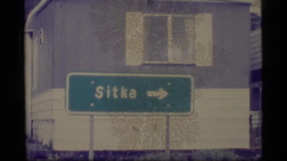1977 Alaska home-movie frame showing the Sitka direction sign from farther back, with building siding and windows behind it.