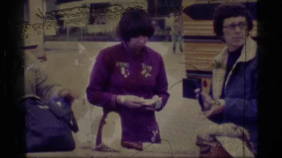 1977 Alaska home-movie frame with two adult women in jackets near bags, one holding small items, in a casual travel scene.