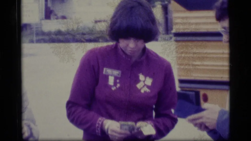 Closer 1977 Alaska home-movie view of a woman in a purple jacket counting money while a nearby hand holds a wallet.