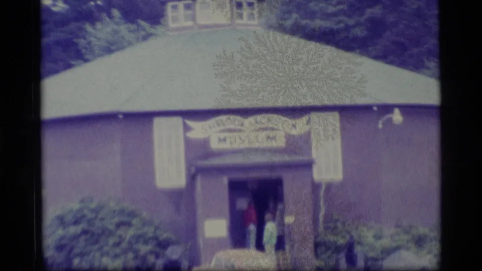 1977 Alaska 8mm view of the Sheldor Jackson Museum building with a curved roof, entrance sign, shrubs, and visitors at the doorway.