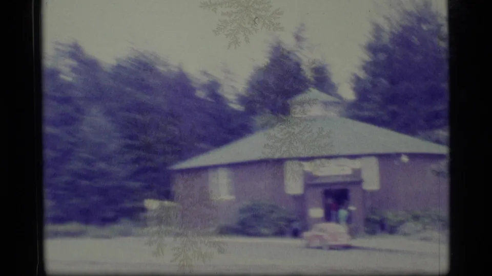Wide 1977 Alaska home-movie frame of the museum exterior surrounded by trees, with a parked car and people near the entrance.