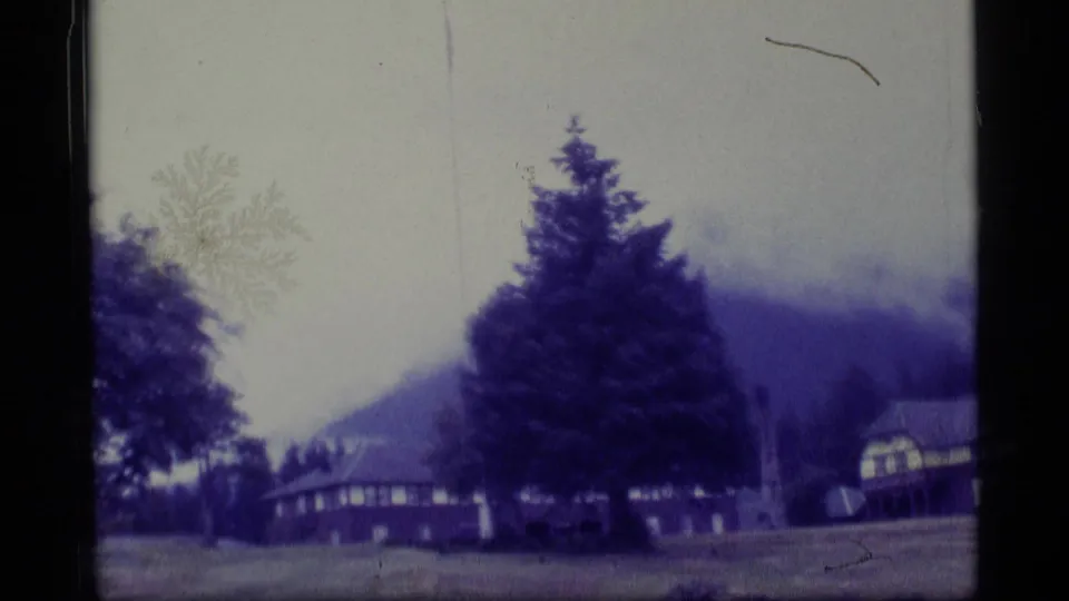 1977 Alaska 8mm still showing a large evergreen, low residential structure, lawn, hillside, and visible film scratches.
