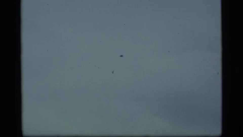 1977 Alaska 8mm frame with a small bird silhouette crossing an overcast blue-gray sky, bordered by dark frame edges.