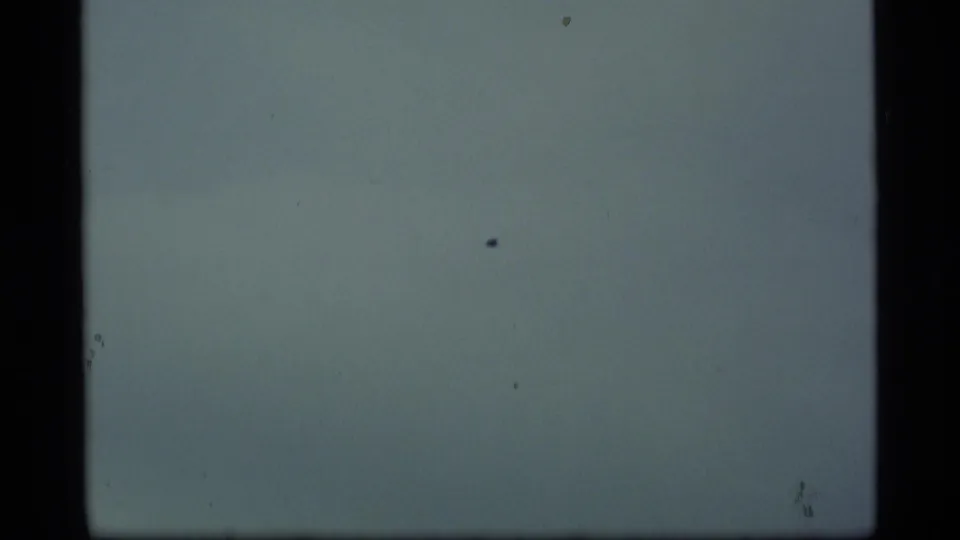 1977 Alaska 8mm frame showing a tiny bird in open cloudy sky, with dark framing and a faint forested horizon at the edge.