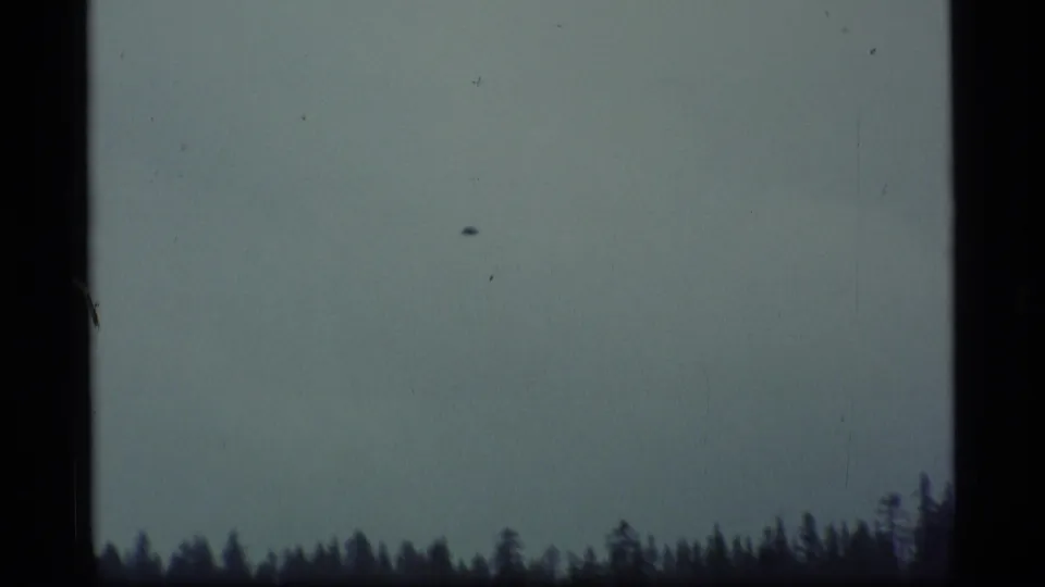 1977 Alaska 8mm frame looking across pale cloudy sky toward a dark forested treeline, with visible film wear and edge framing.