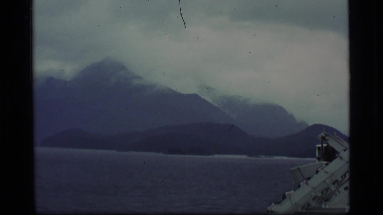 1977 Alaska 8mm frame showing dark water, layered mountains, low cloud, and part of a white vessel railing.