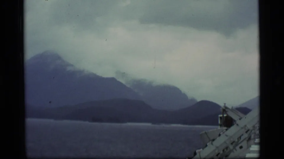 Alaska home-movie view across water toward cloud-wrapped mountains, framed by a white vessel railing.