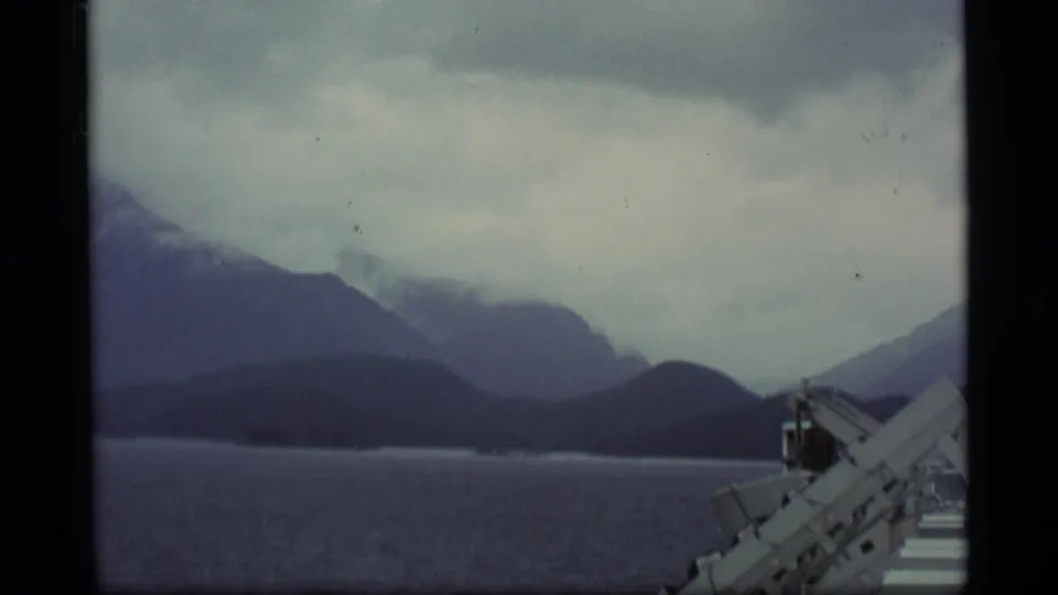 Vintage 8mm Alaska landscape from a moving vessel, showing water, misty mountains, and white railing detail.