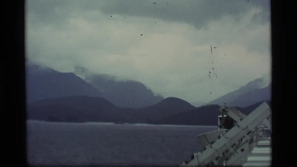 1977 Alaska 8mm still with layered mountain silhouettes, overcast sky, water, and foreground ship railing.