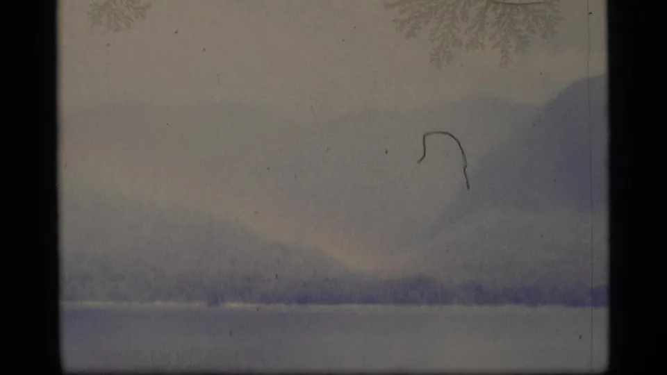 1977 Alaska 8mm frame showing blue-gray mountain forms, a pale waterline, and visible dust or scratch marks.