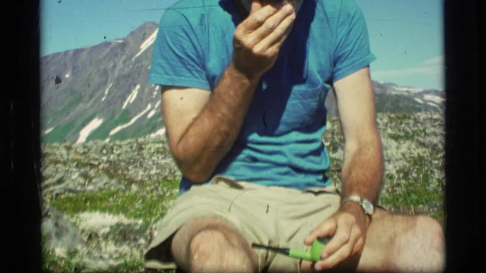 In Alaska 1977 home-movie footage, a seated man in a blue T-shirt and khaki shorts holds a green pipe before distant mountains.