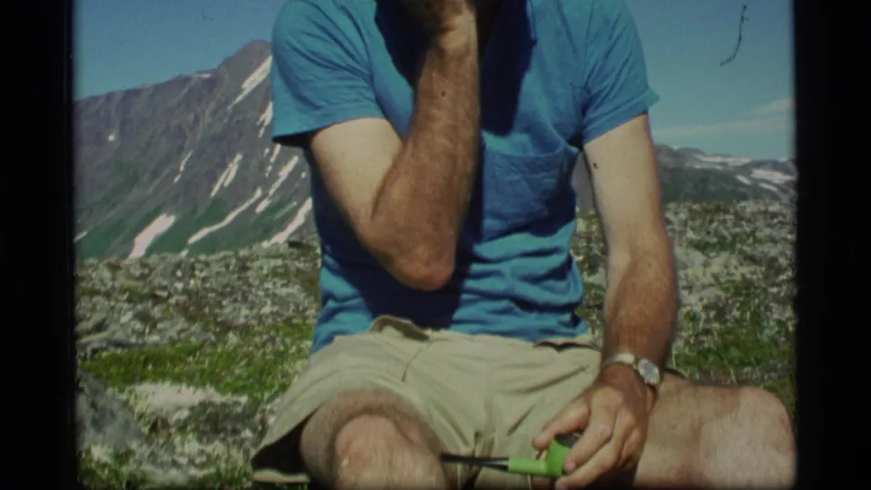 In Alaska 1977 home-movie footage, a seated man holds a green pipe as rugged mountains and snow patches appear behind him.