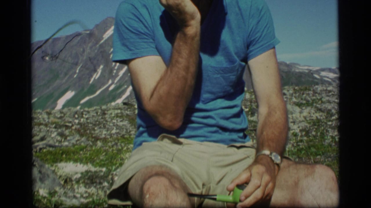 In 1977 Alaska footage, a seated adult man holds a green pipe with snow-streaked mountains and clear sky behind him.