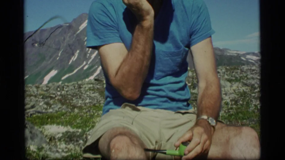 In 1977 Alaska footage, a seated adult man holds a green pipe with snow-streaked mountains and clear sky behind him.