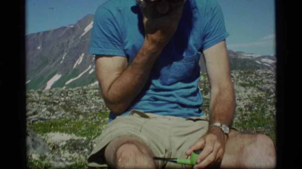 A home-movie frame from Alaska shows the seated man rubbing his beard, pipe in hand, with alpine terrain behind him.